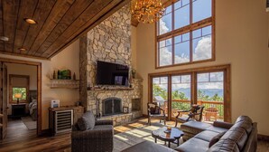A spacious living room with a stone fireplace, leather sofas, and large windows offering a view of the outdoors. - Majestic View Hideaway at Eagles Nest (Banner Elk)