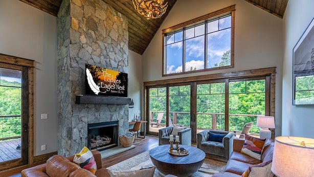A spacious living room with a stone fireplace, leather sofas, and large windows offering a view of the outdoors. - Olive and Georgia's at Eagles Nest (Elk Park)