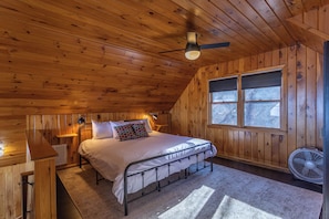 Une chambre dans un chalet en bois, avec un lit, un ventilateur de plafond, une fenêtre et un ventilateur posé sur le sol.