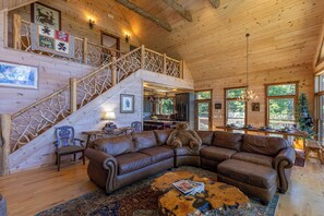 Interior de una cabaña de madera con un amplio sofá seccional de cuero marrón, una escalera de madera rústica y un comedor con un árbol de Navidad.