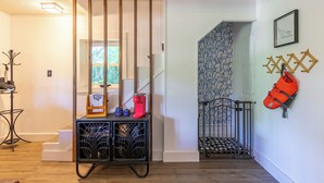 Un couloir avec un escalier, un porte-manteau sur lequel est accroché un sac rouge, un présentoir métallique noir avec divers objets, et un mur à motifs.