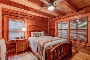 Une chambre dans une cabane en bois, avec un lit, des tables de chevet, une lampe et un ventilateur de plafond.
