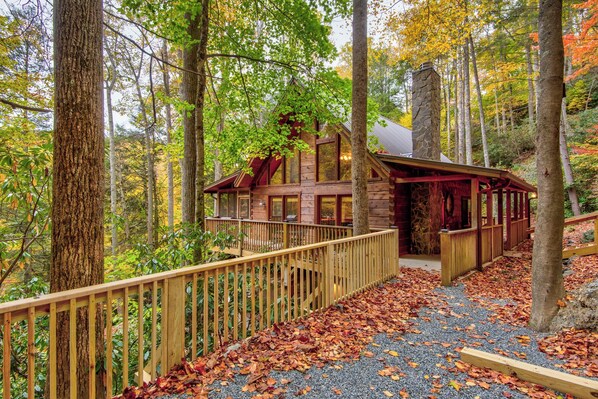 Une cabane dotée d’une terrasse en bois et d’une cheminée en pierre, entourée d’arbres et de feuilles d’automne.