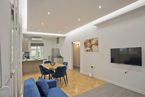 A modern living room with a sofa, a dining table with chairs, a TV mounted on the wall, and a kitchen area visible through an open door. - Like Home in Kalithea (Kallithea)