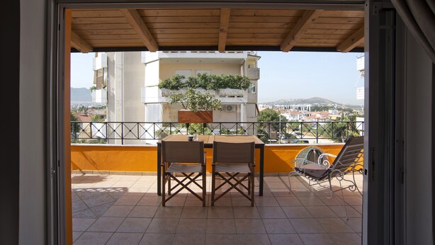 A balcony with a wooden table and chairs, a chair with a cushion, and a view of a cityscape with buildings and mountains in the background. - Apos Place in Pefki With 1 Bedrooms and 1 Bathrooms (Lykovrysi-Pefki)