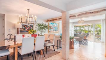 A spacious dining area with a glass table, white chairs, and a chandelier. There is a large painting on the wall and a view of the outdoors through large windows.
