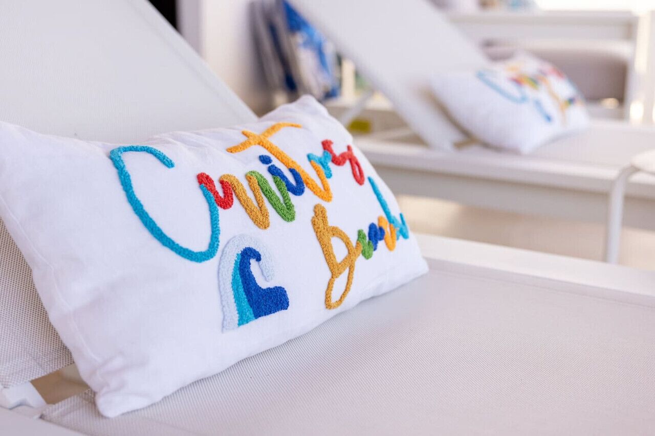 A white lounge chair with a colorful embroidered pillow that reads "Cunning & Brave."