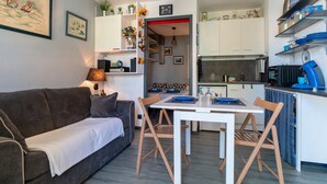 A compact living space with a sofa, dining table, and chairs. A kitchen area with cabinets and appliances is visible. - Le P'tit Loup de Mer - Appt proche plage, terrasse (Saint-Nic)