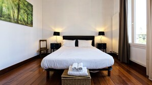 A neatly arranged hotel room with a large bed, two bedside tables, a nightstand, a wicker bench, and a painting on the wall.