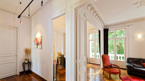 A spacious living room with hardwood flooring, a red armchair, a black sofa, and a large window with curtains.