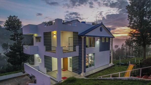 Una casa moderna de dos pisos con balcón, una escalera y un tobogán, rodeada de vegetación.