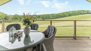 A round table with a glass top, surrounded by wicker chairs with grey cushions, set on a wooden deck with a view of a green field and trees. - Safari Tent Two - UK44405 (Ross-on-Wye)