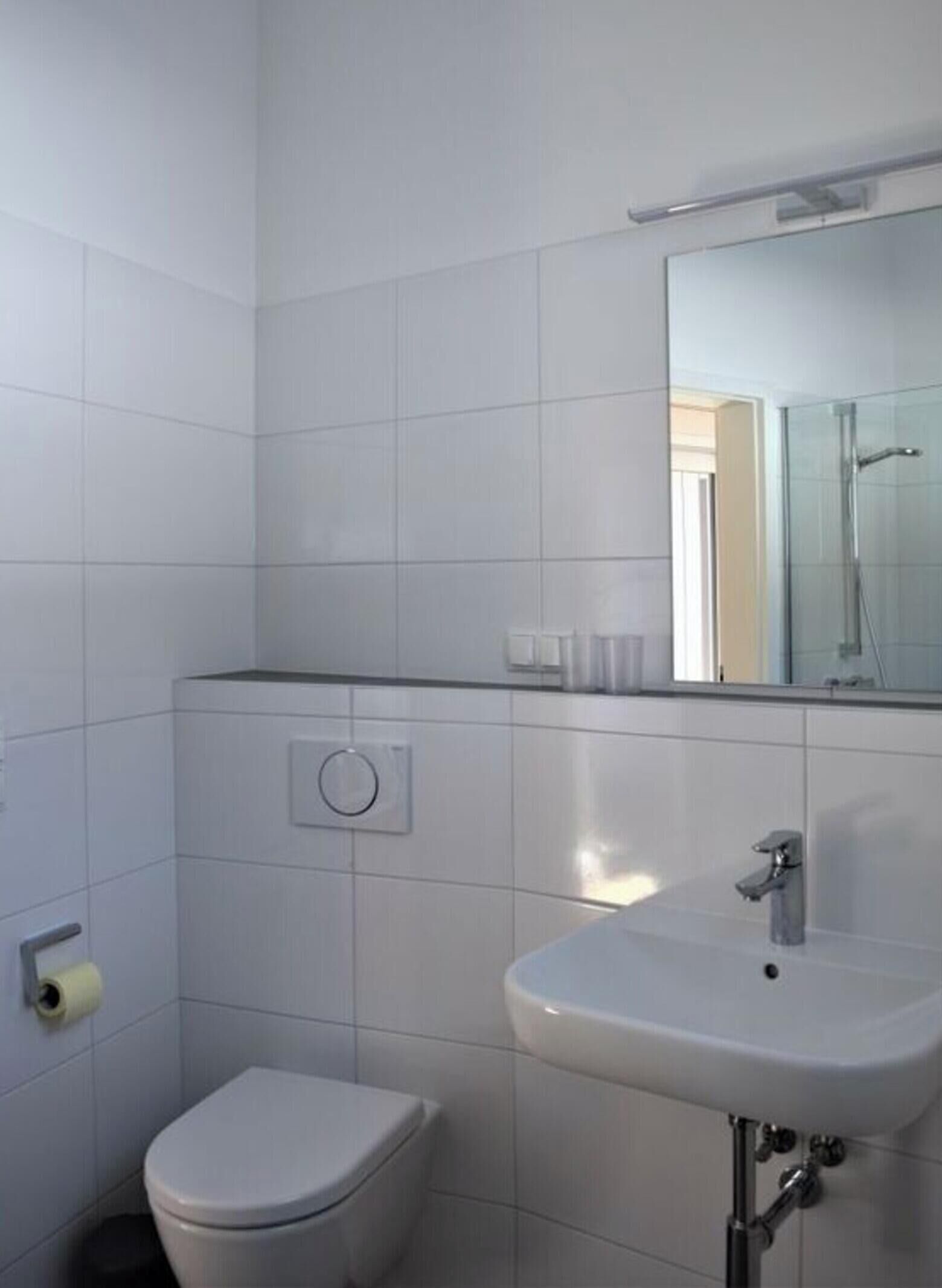 A small, clean bathroom with white tiles, a toilet, a sink, and a mirror.