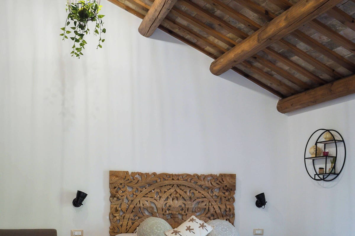 A bedroom with a wooden ceiling, a bed with a decorative headboard, a nightstand, a wall-mounted shelf, and a geometric-patterned floor.