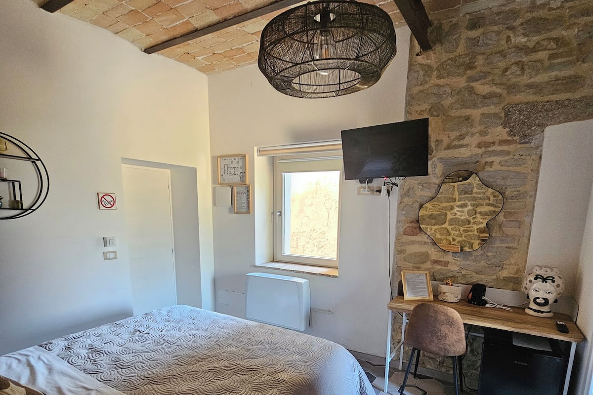 A bedroom with a large bed, a desk with a computer, a TV, and a stone wall.