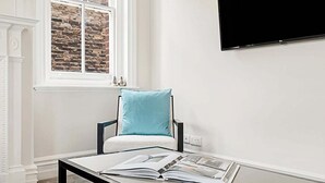 Quarto com parede branca, janela com padrão de tijolos, cadeira preta com almofada azul clara, mesa com uma revista aberta e televisão na parede.
