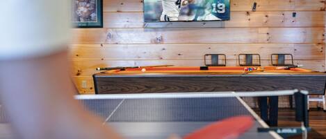 Una mesa de billar con una vara de billar en primer plano, un televisor montado en una pared de madera mostrando imágenes deportivas, y un cuadro enmarcado a la izquierda.