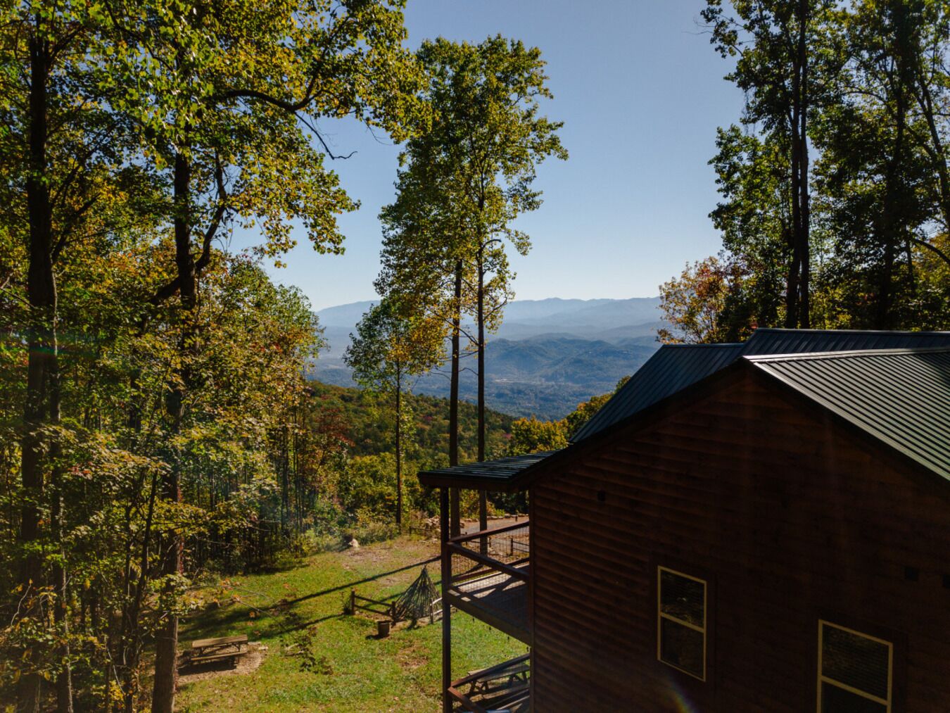 Una cabaña con techo de metal y terraza de madera, rodeada de árboles y un paisaje montañoso.