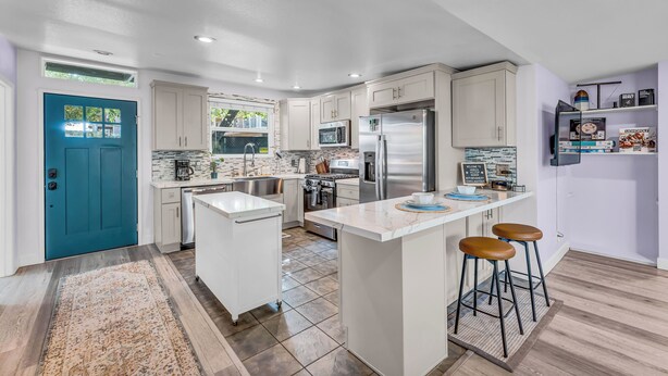 A modern kitchen with white cabinets, a central island with stools, stainless steel appliances, and a teal door.