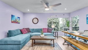 A modern living room with a teal sectional sofa, a wooden coffee table, and a dining area with a wooden bench and table.