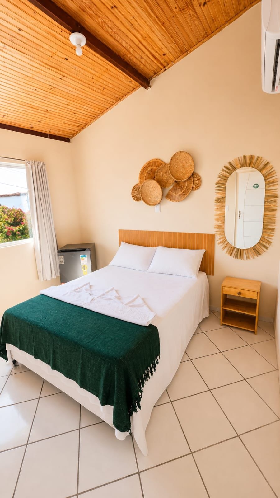 A bedroom with a bed, a wooden ceiling, a wall-mounted mirror, and a window with curtains.