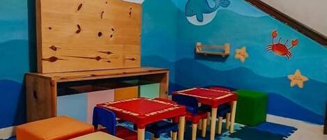 Un cuarto de juegos para niños con un muro decorado con motivos marinos azules, mesas de juego de madera y asientos coloridos.