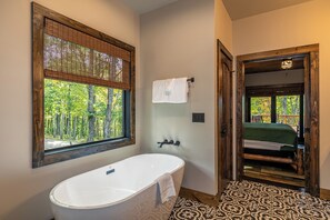 A bathroom with a freestanding bathtub, a window with a view of trees, and a door leading to a bedroom with a bed and a balcony. - Beagles Rest at Eagles Nest (Banner Elk)