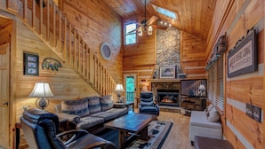 A log cabin interior with a fireplace, leather seating, a stone accent wall, and a ceiling fan. - Carolina Charm (Banner Elk)