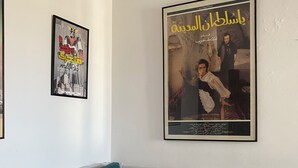 Ein Wohnzimmer mit grauem Sofa und blaumetallenen Kissen, einem gerahmten Filmplakat an der Wand und einem gerahmten Bild über dem Sofa.