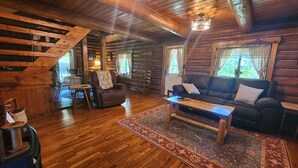 Interior de una cabaña de troncos con piso de madera, un sofá, una mesa de centro y una escalera que sube a un nivel superior.