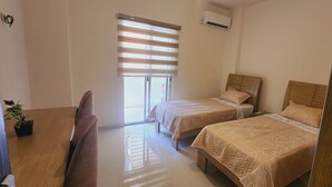 A bedroom with two beds, a window with blinds, an air conditioning unit, and a potted plant.