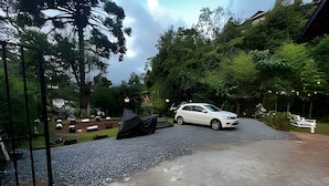 A white car parked on a gravel driveway in a lush, green area with a metal gate on the left. - Fracalanza (Campos do Jordão)