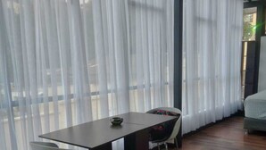 A room with a wooden table and two chairs, large windows with sheer curtains, and a view of the outdoors.