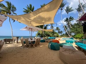 A beachside lounge with seating and tables under a canopy, surrounded by palm trees and the ocean. - Beautiful 1BR in front of the beach with pool spa restaurant (Las Terrenas)