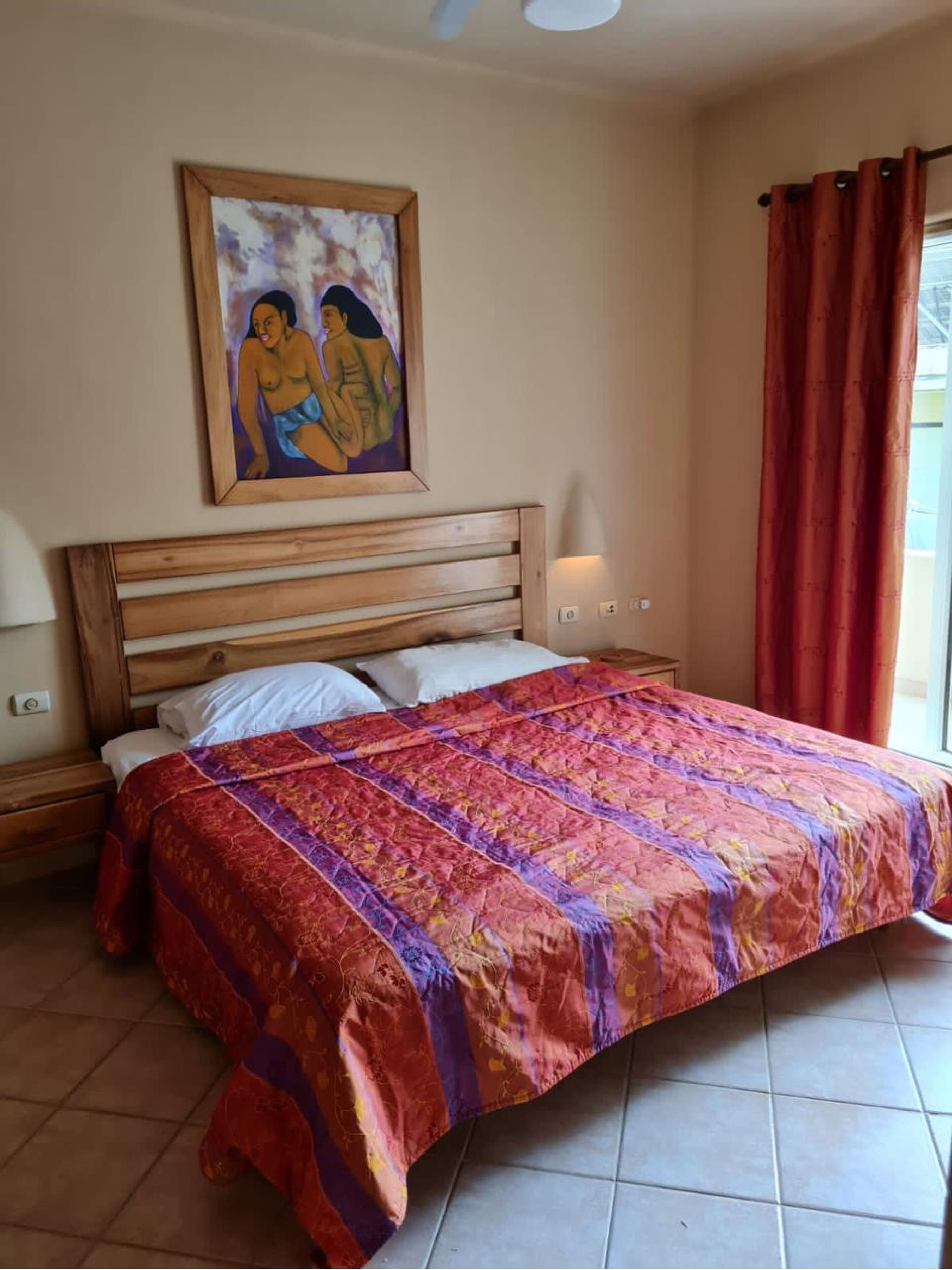 A bedroom with a wooden headboard, a large bed with a colorful quilt, a painting on the wall, and a window with red curtains.