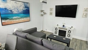Ein Wohnzimmer mit grauem Sofa, einem flachbildfernseher an der Wand, einem Kamin mit weißem Schrank und einem gerahmten Bild von einer Strandansicht.