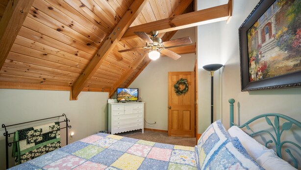 A bedroom with a wooden ceiling, a bed with a colorful quilt, a dresser, a television, a door with a wreath, and a painting on the wall. - Appalachian Satisfaction (Banner Elk)