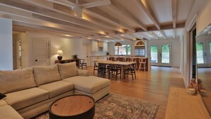 A spacious living area with a large sofa, a round coffee table, and a dining table with chairs. There is a ceiling fan and recessed lighting.