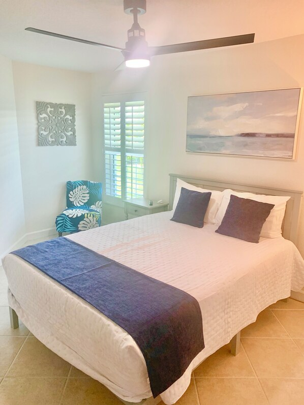 A neatly made bed with a white and blue bedspread, a ceiling fan, a window with blinds, and a painting on the wall. - SENSATIONAL!! WATER-VIEW, LUXURY REMODEL, SUNNY HEATED POOL, HUGE 145’ SEA WALL (Punta Gorda)