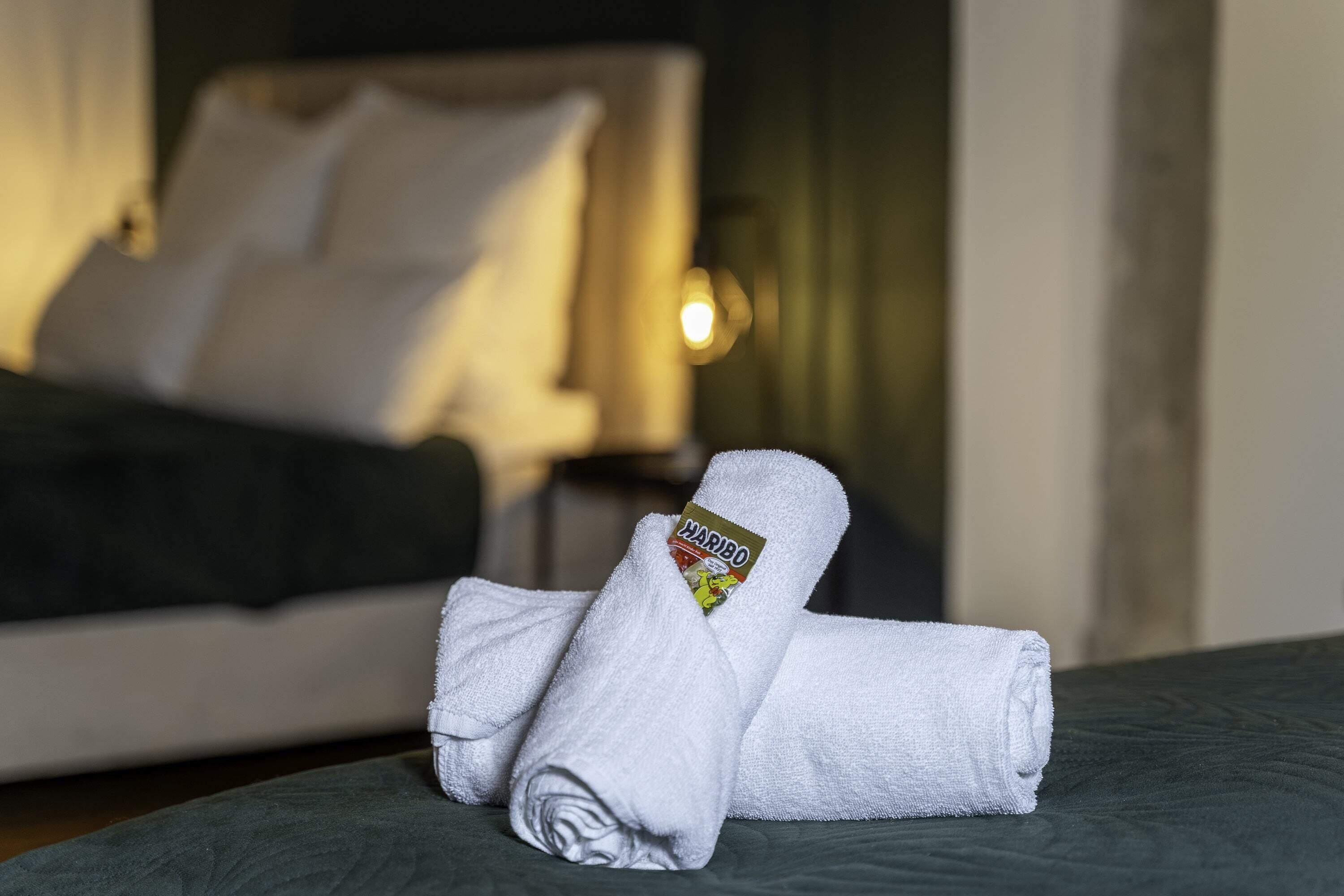 A neatly made bed with a white towel and a Haribo logo, a dark green bedspread, and a white pillow in a hotel room.