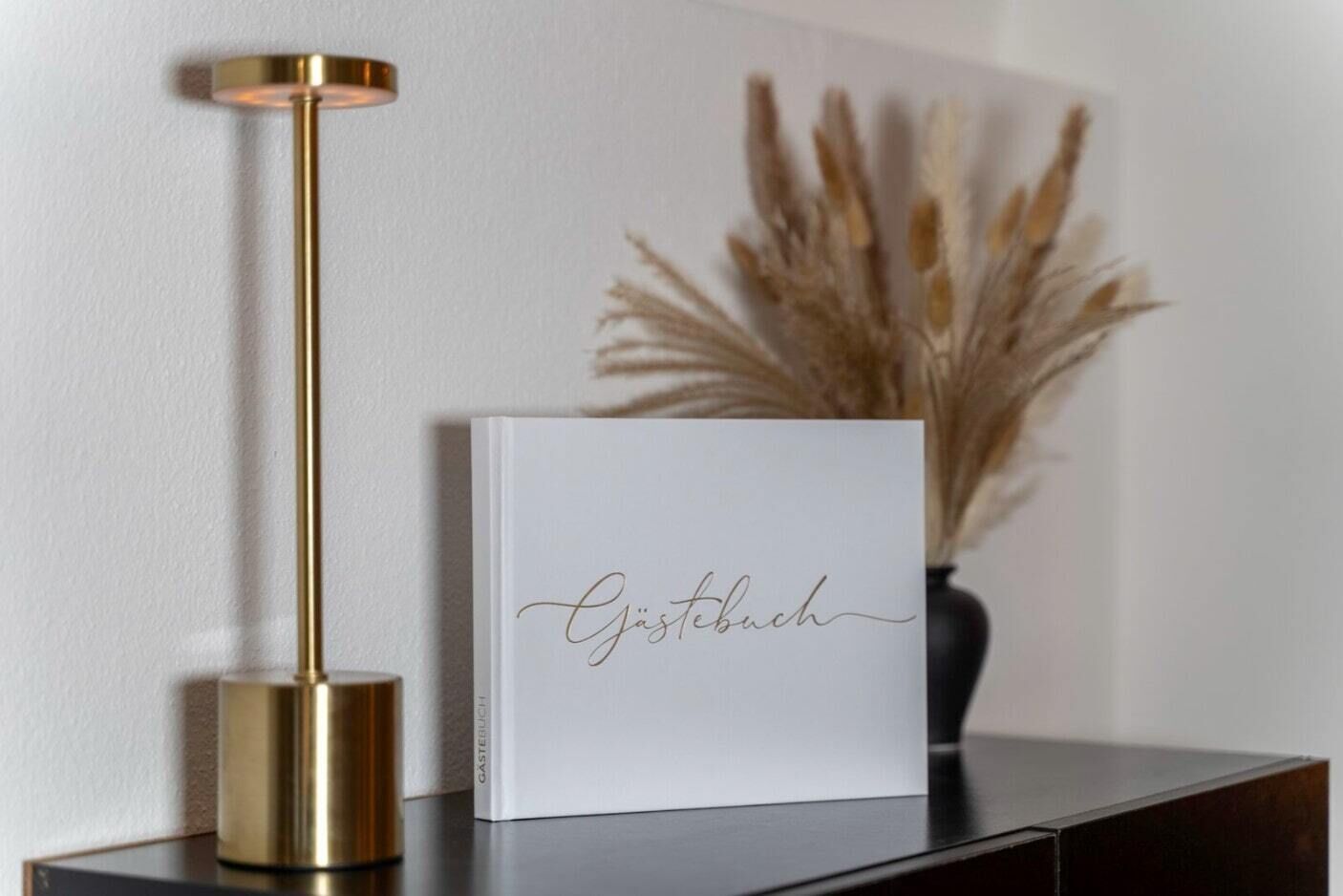 A white framed book with the word "Gästebuch" on it, placed on a dark surface next to a gold-colored floor lamp.