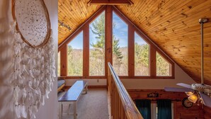 Une pièce dotée d’un plafond en bois incliné, de grandes fenêtres, d’un attrape-rêves, d’un banc en bois et d’un ventilateur de plafond.