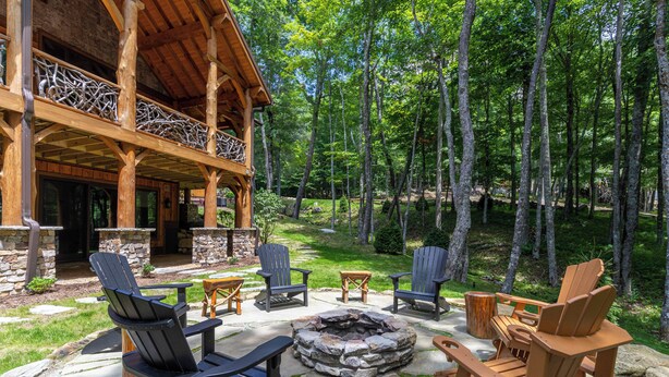Un patio al aire libre con sillas Adirondack, un hogar de piedra y una terraza de madera con vista a una zona boscosa.