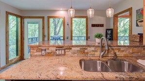 A kitchen with granite countertops, a stainless steel sink, hanging pendant lights, and a view of the outdoors through large windows. - Paws A While (Blowing Rock)