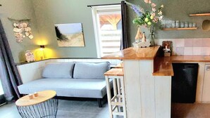 A compact living space with a sofa, a small round table, a kitchen area with a dishwasher, and a window with curtains.
