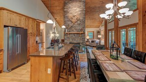 A spacious kitchen with wooden cabinets, a large island with stools, a stone fireplace, and a dining area with a long table and chairs. - High On Cloud Nine (Boone)