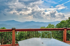 A table with a metal top, surrounded by a wooden railing, overlooking a mountainous landscape with trees and a cloudy sky. - Shooting Star Lodge (Boone)