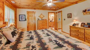 A cozy bedroom with a wooden ceiling, a bed with a forest-themed quilt, a wooden dresser, and a ceiling fan. - Kaylee's Kabin (Vilas)