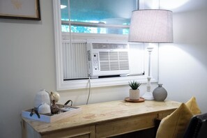 Una habitación con ventana, aire acondicionado, lámpara, planta pequeña y un florero sobre una mesa.