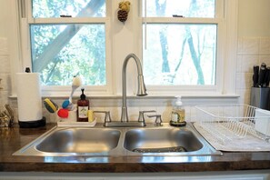 Une cuisine équipée d’un évier à deux bacs, d’une fenêtre donnant sur les arbres et de divers ustensiles de cuisine disposés sur le plan de travail.